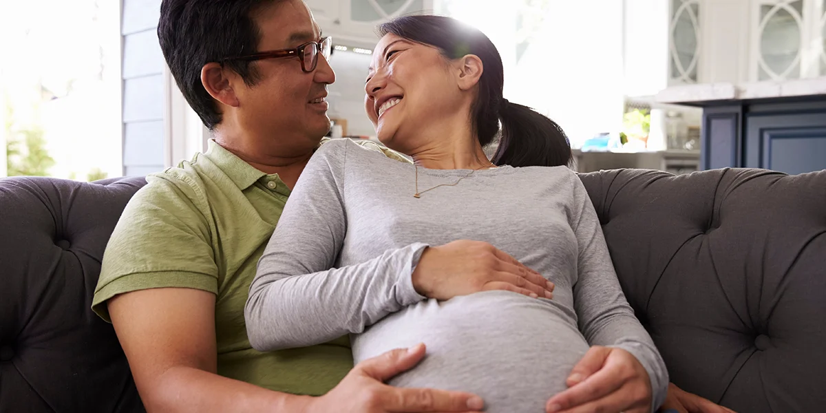 Happy asian couple touching their pregnant belly while relaxing on a couch