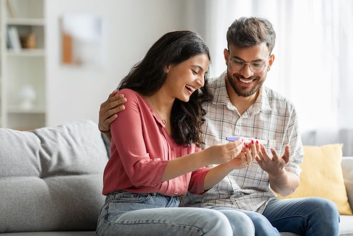 Happy indian spouses holding positive pregnancy test, sitting on couch at home together, couple celebrating awaiting baby, copy space