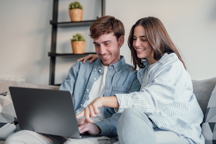 Happy millennial couple sit relax on couch in living room watching video on laptop together, smiling young husband and wife rest on sofa at home browsing Internet using modern computer device