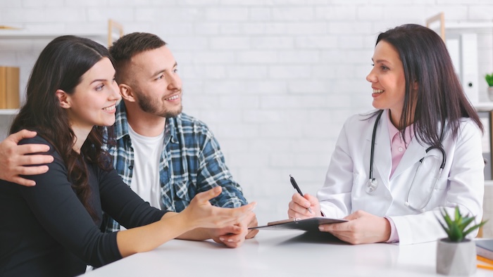 European couple sits at a table with woman egg donation doctor in a white coat. The couple is engaged in a conversation, listening intently as the doctor takes notes
